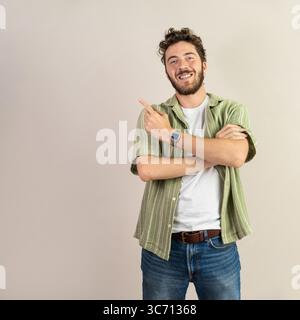A confident and friendly young man with a beard smiles at the camera and points to his side, presenting an empty space on a neutral background for an Stock Photo