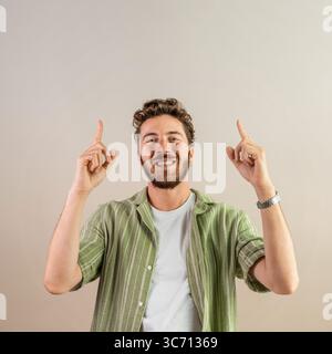 A cheerful smiling young man with a beard looks at the camera and points upwards with both hands, isolated on a beige background with ample copy space Stock Photo