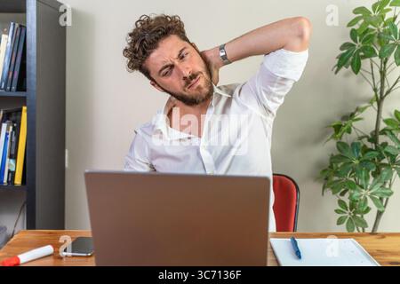 A tired young businessman or student feels a sharp pain in his stiff neck, a result of poor posture and long hours of sitting at a desk with a compute Stock Photo