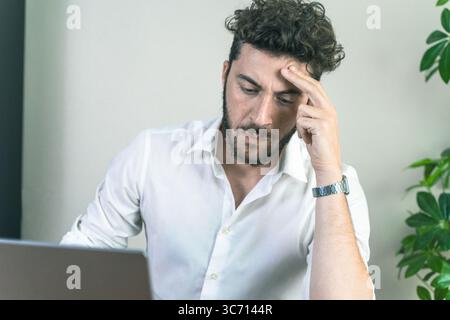 A stressed and tired young businessman touches his temple, feeling the pain of a headache or eye strain after concentrating on his computer for too lo Stock Photo