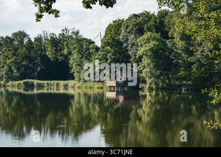 A peaceful view of a small wooden cabin built on stilts over a calm forest lake in Kreuzlingen, Switzerland. Surrounded by dense green trees and refle Stock Photo