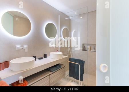 Modern bathroom featuring twin sinks, round LED mirrors, and walk-in glass shower in neutral design. Stock Photo