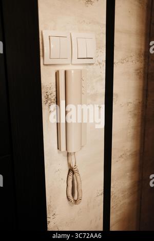 Wall-mounted intercom telephone with coiled cord next to two light switches on a textured wall in hallway. Stock Photo