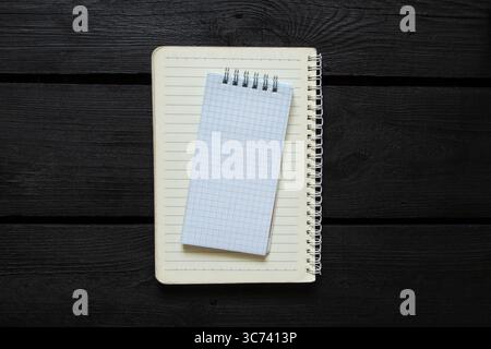 Open notebook in line with white checkered notebook on top, both on spiral, lying on black wooden background and top view, template for notes Stock Photo
