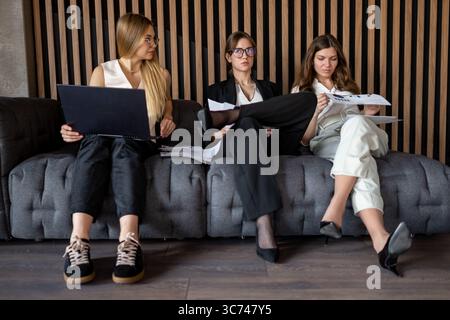 Businesswomen reviewing project data on paper and laptop while seated on office couch Stock Photo
