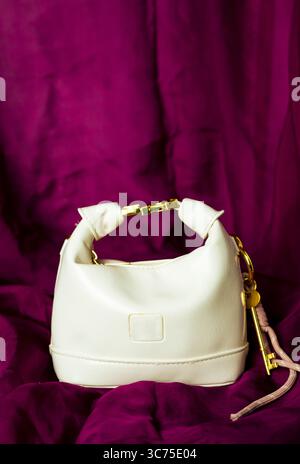 Close-up view of a gold-toned key charm attached to a white leather purse, resting on a rich purple fabric background. The key charm features decorati Stock Photo