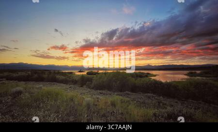 A vibrant sunset paints the sky with fiery hues over a tranquil lake, with silhouetted mountains in the background. Stock Photo