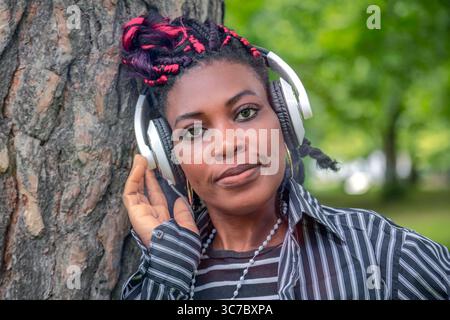 Beautiful African woman enjoying music with headphones while leaning against tree in park. Peaceful portrait with hand touching headphones wearing str Stock Photo
