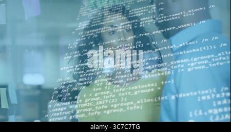 Reviewing team in green and blue shirts standing behind clear code screen at office, with monitors Stock Photo