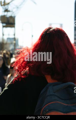 Woman with vibrant, flowing red hair walks down a bustling city street, her backpack slung over her shoulder Stock Photo