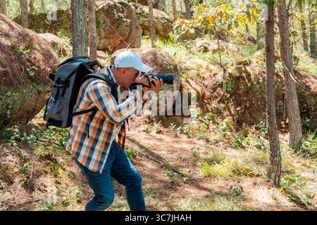 Latin American male photographer crouching with camera to frame the perfect shot in a forest. Outdoor adventure photography scene with natural light a Stock Photo