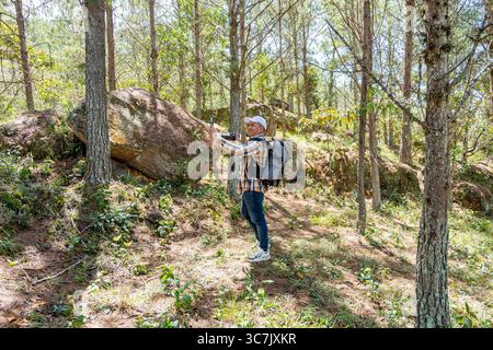 Photographer in a pine forest smiling as he takes a selfie with one hand and holds his camera in the other, capturing the behind-the-scenes of his adv Stock Photo