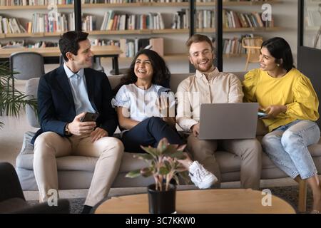 Four colleagues holding diverse devices talking seated on sofa Stock Photo