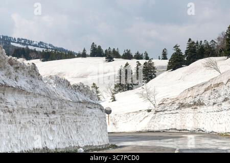 Snow wall lines the side of National Route 103 from Aomori, Japan, towards Mount Hakkoda in early spring with snow covered mountains in background Stock Photo