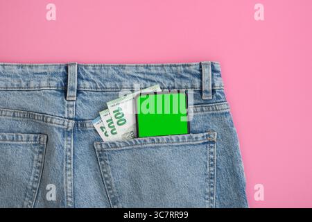 Smartphone green screen mockup and euro bills in back pocket of blue jeans on pink background. Banner with no people. Stock Photo