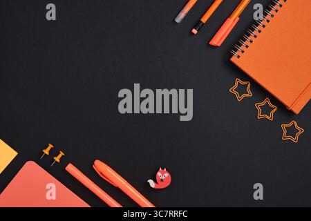 Stationery background with orange pencil, pen, marker, clips, notebook on black backdrop Stock Photo