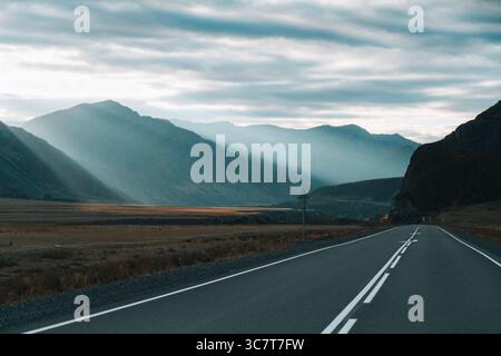 A wide view of a winding road with a guardrail leading toward distant mountains with a dramatic cloudy sky and sunrays breaking through Stock Photo