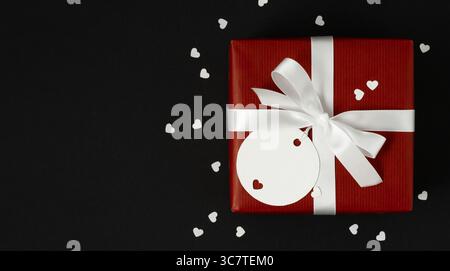 Red gift box with white bow and empty white round gift tag with heart. Space for text. Black background. Valentine's Day Card Stock Photo