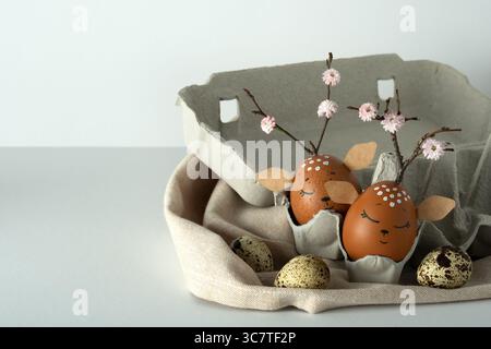Decorated eggs with deer faces and blooming twigs in an egg carton on fabric napkin. Concept of Easter creativity and spring holiday crafts. Stock Photo