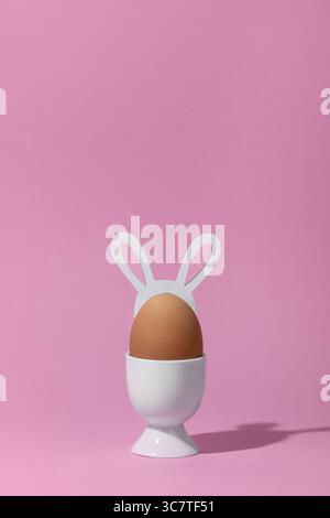 Brown egg with hare ears in a white eggcup on pink background. Minimal easter concept. Stock Photo