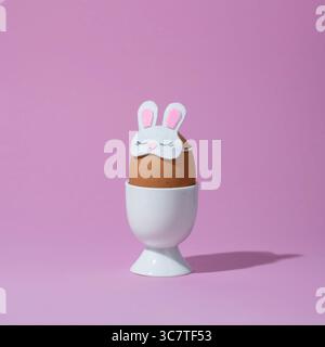 Brown egg in bunny sleep mask in white egg cup on pink background. Easter egg concept. Stock Photo