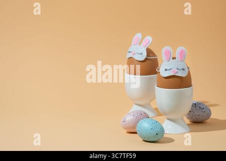 Brown eggs in bunny sleep masks in white egg cups on pastel yellow background. Easter banner, space for your text. Stock Photo