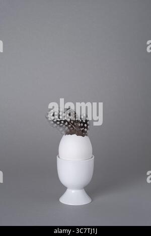 White eggcup with vase of white eggshells and guinea fowl feathers on grey background. Easter concept, copyspace. Stock Photo