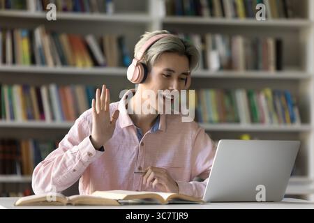 Smiling guy student wearing headphones waving hand to laptop screen Stock Photo