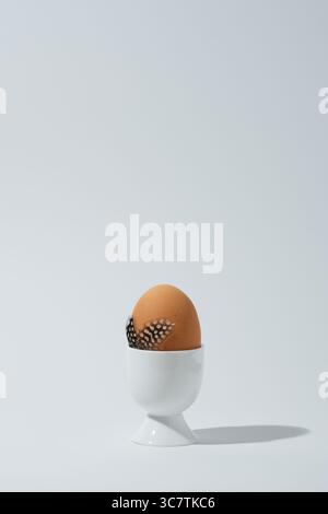 Brown egg in white eggcup with guinea fowl feathers. White background, space for text. Easter card. Stock Photo