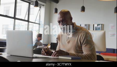 Adult man wearing beige sweater working on silver laptop in shared office, with pen and notebook Stock Photo