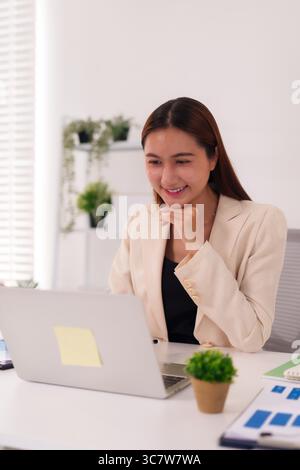 Adult Asian professional female happily working on computer in a clean workspace focused on business tasks and online solutions. Stock Photo