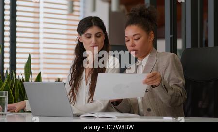 African American Caucasian businesswomen two female coworkers multiethnic business partners colleagues girls women together discuss papers graphs Stock Photo