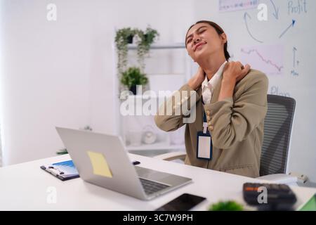 Adult Asian woman experiencing discomfort and pain in her neck and shoulders from long hours working at her office desk seeking relief Stock Photo
