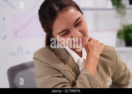 Adult Asian woman in an office suffering from intense neck and shoulder discomfort from prolonged computer work stress and fatigue Stock Photo