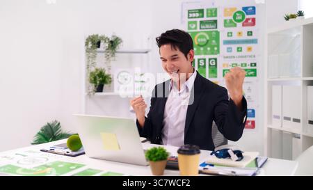 Adult Asian man jubilant celebrating professional success in office environment looking at laptop screen achieves goals Stock Photo
