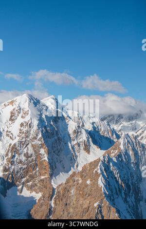 A dramatic vertical view of a rugged, snowcapped mountain range under a clear blue sky with some soft clouds in the distance Stock Photo