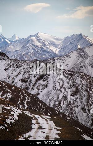 A rugged, snow-covered mountain with a winding road leading towards a majestic snowcapped mountain range under a cloudy sky Stock Photo
