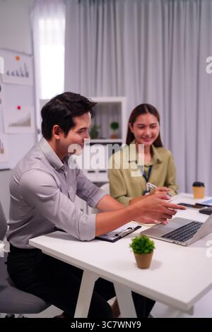 Two Asian business professionals collaborate on a laptop discussing strategies and solutions in a modern office environment Stock Photo