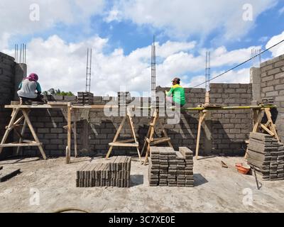 Badung, Bali, Indonesia – July 13, 2025: Construction workers teamwork in building a concrete block wall with wooden scaffolding and masonry tools Stock Photo