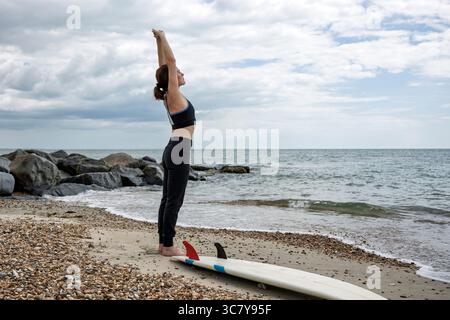 woman surfer doing stretching exercises on the beach by her surfboard Stock Photo