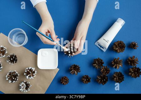 Person painting pine cones Christmas on a blue background. Preparation of Christmas decorations. Stock Photo