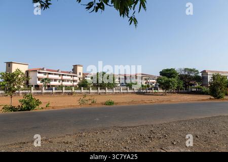 A rural asphalt road stretches into the distance, flanked by dry grass and scattered trees under a clear blue sky. Indian rural road with building. Stock Photo