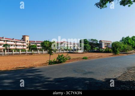 A rural asphalt road stretches into the distance, flanked by dry grass and scattered trees under a clear blue sky. Indian rural road with building. Stock Photo