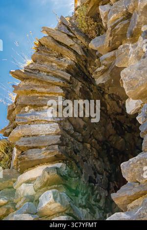 Old stone arch made of stacked slabs with sharp edges, part of historic ruin on rocky slope under blue sky and vegetation. Stock Photo