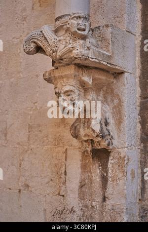 Weathered limestone sculptural capital featuring carved human faces and figures, showcasing medieval craftsmanship on historic building Stock Photo