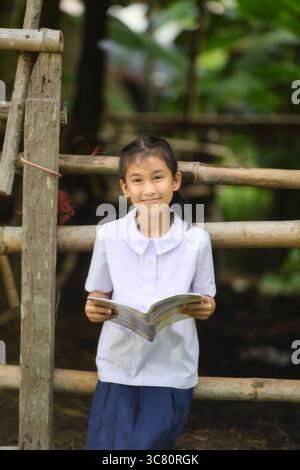 Portrait of a Smiling girl wearing school uniform sitting on a wooden fence reading a book, Thailand Stock Photo