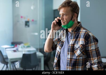 Man holding smartphone wearing green face mask in office by sticky note partition, copy space Stock Photo