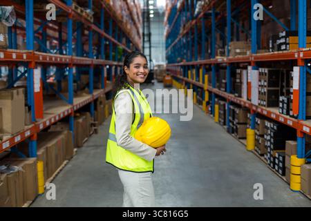 Warehouse manager wearing reflective vest and holding yellow hard hat among shelving with boxes Stock Photo