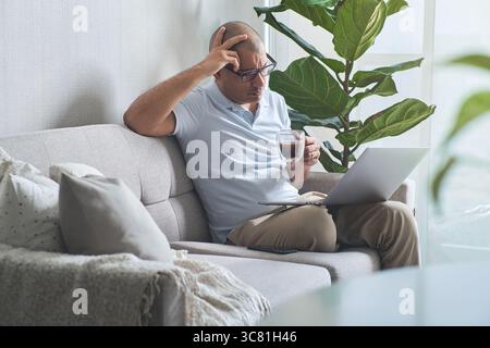 Concentrated businessman drinking coffee and using laptop while working remotely at home Stock Photo