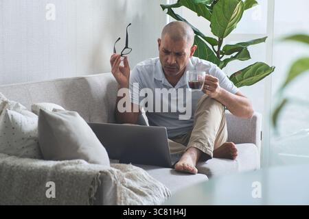 Concentrated businessman drinking coffee and using laptop while working remotely at home Stock Photo